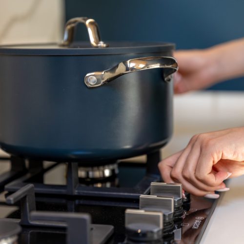 A close view shows a hand rotating a gas stove control beside a dark lidded pot. Crisp detail signals the start of cooking and a warm kitchen mood.